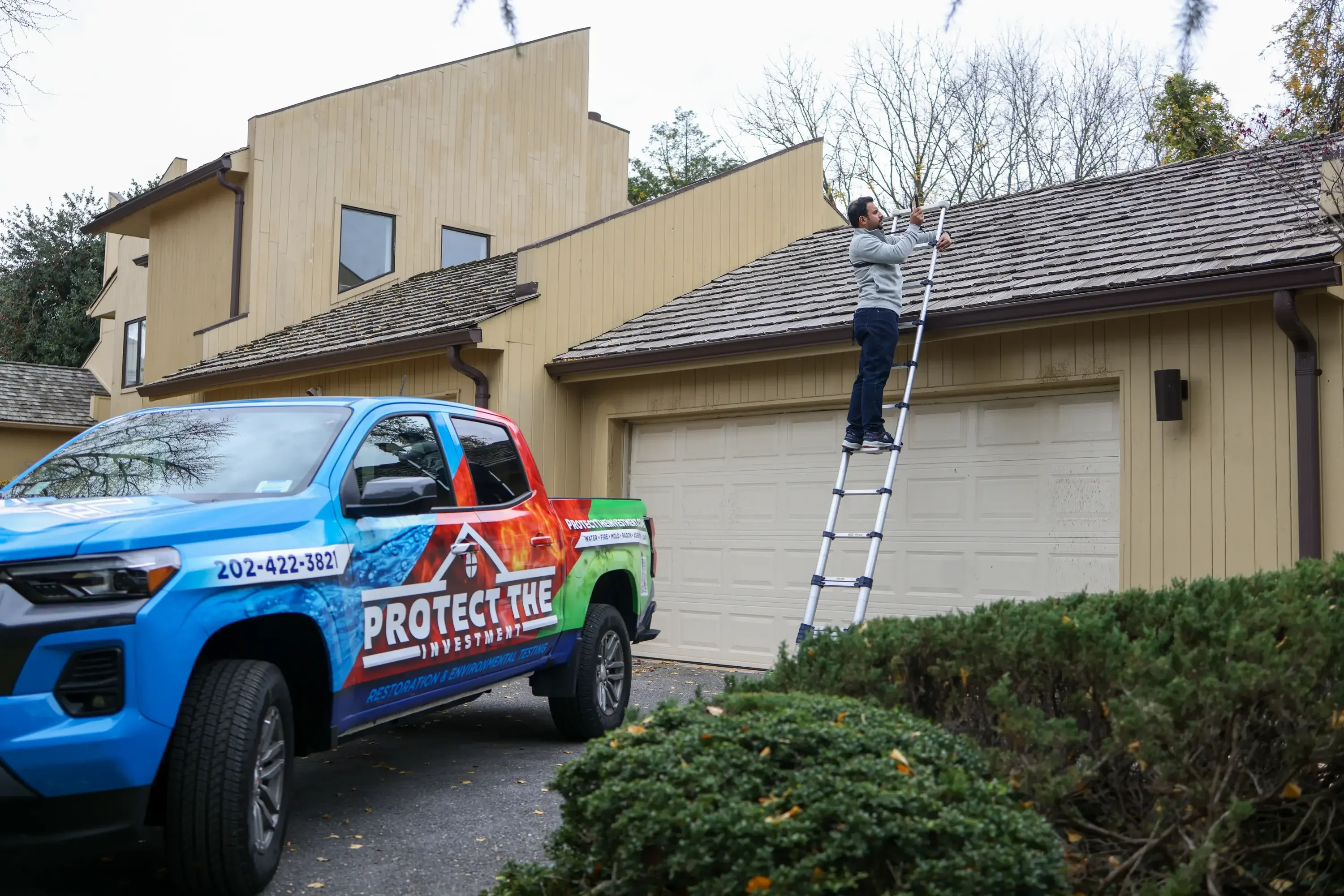 Home inspector on ladder inspecting roof of tan metal-sided house with garage